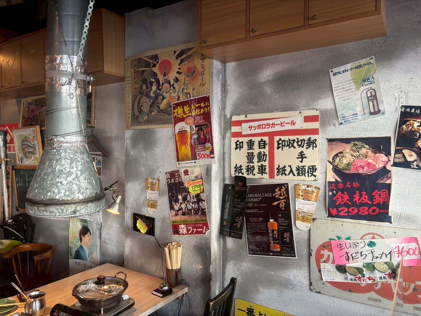 店舗内装。昭和レトロなポスターや看板が壁一面に飾られている。テーブル上には鍋と換気ダクトがあり、賑やかな雰囲気が漂っている。